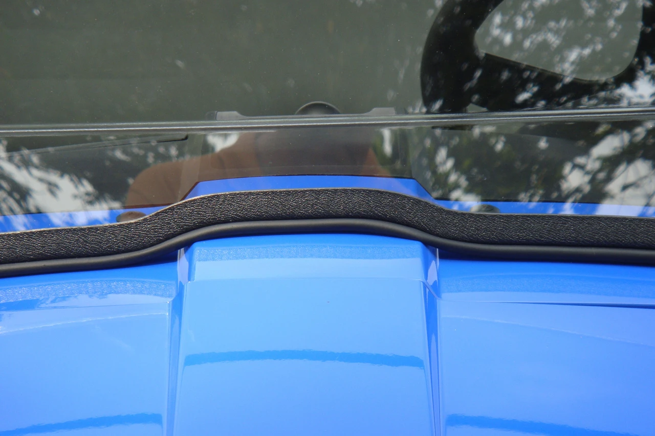 Close-up of the 3 Star two-piece windshield lower edge with black rubber seal on a blue New Holland Rustler 115 hood