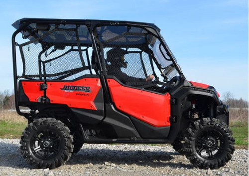 Side profile of the red Honda Pioneer 1000-5 on gravel showing the SuperATV tinted roof over the cage, driver seated inside