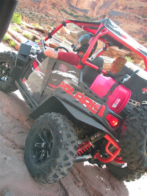 Rear three-quarter view of the Polaris RZR with Trail Armor doors and a helmeted rider on red rock, portrait crop