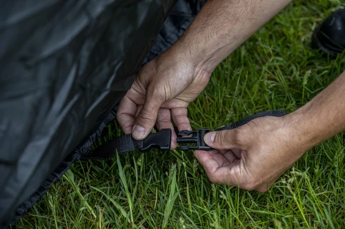 Hands clipping the buckle strap of the Kolpin mid-size UTV cover at the bottom edge on grass