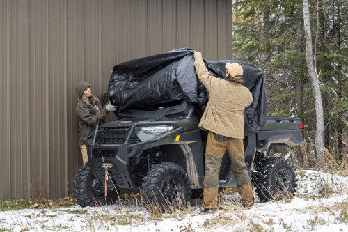 Two people pulling the Kolpin black UTV cover over a side-by-side beside a metal building in the snow