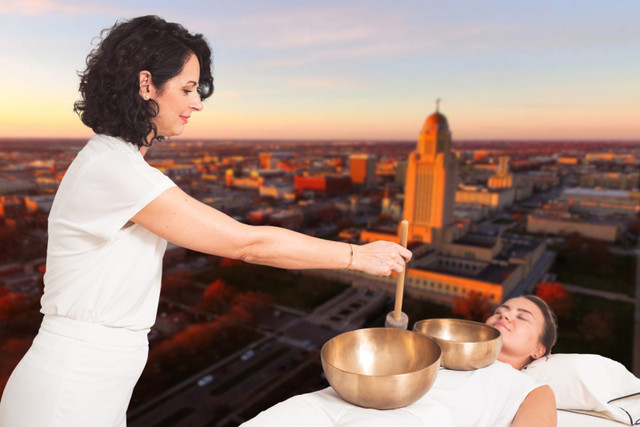 Vibrational Sound Therapy practitioner playing singing bowls for a client on a massage table, Lincoln, NE skyline