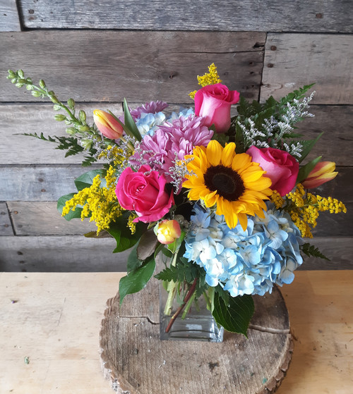 Blue hydrangea, yellow sunflower, pink roses, golden solidago, purple daises, arranged in a glass vase with seasonal greenery.