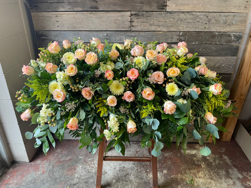 A casket blanket filled with soft yellows, peaches, and a variety of pink roses and filler