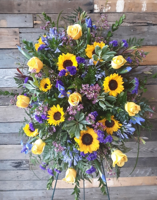 A compact spray filled with greenery, purple filler, purple iris, yellow roses and sunflowers.
