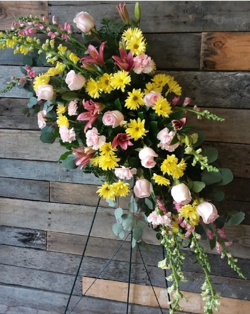An angled teardrop shaped standing spray filled with a variety of yellows and soft pink flowers. This arrangement includes daisies, roses, carnations, and complimenting greenery.