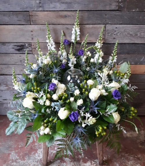 An arrangement to surround an urn on all sides, higher in the back and low in the front, filled with greenery, white roses, purple lisianthus, white snapdragons, blue thistle, and seasonal accents.