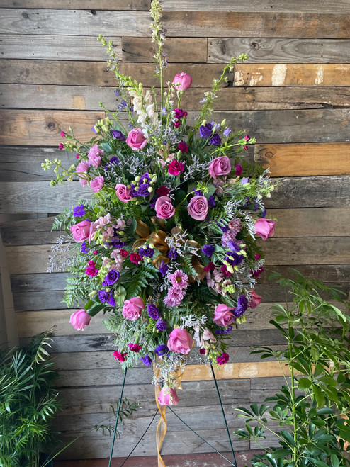 Standing spray with purple roses, purple lisianthus, deep pink carnations, snapdragons, and seasonal greenery.