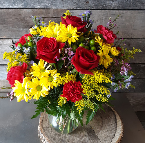 A vibrant, colorful arrangement with yellow daises, red roses, hypericum berries, with greenery in a glass vase.