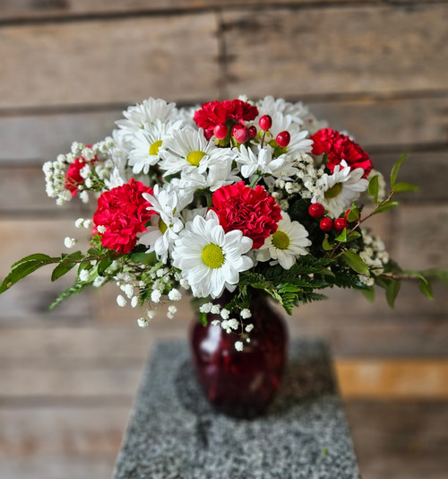 Red carnations, white daisies and delicate baby's breath, with greenery in a ruby red vase.