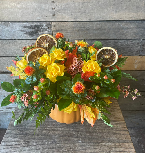 Yellow roses, orange safflower, hypericum berries, wax flowers, seasonal greenery, fall leaves, with dried orange slices, arranged in a pumpkin-shaped container.