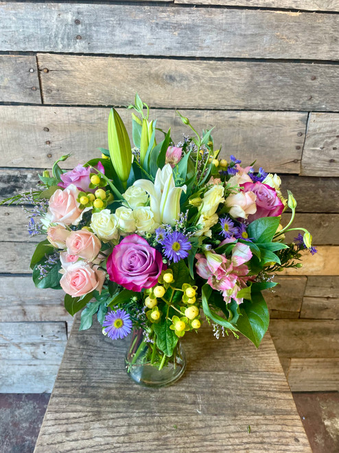  Pink and lavender roses paired with elegant white lilies, creamy lisianthus, pink alstroemeria, lavender asters, yellow hypericum berries, and seasonal accents in a clear glass vase.