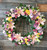 A fresh floral wreath on a stand that is packed with light colors and lush greenery. This showcases lilies, carnations, and mums all in complementing shades of lavenders, pinks and yellows.