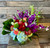 Red roses with vibrant purple orchids and stock, soft blue hydrangea, coral roses, green dianthus, white veronica, and striking pincushion protea with greenery in a glass vase.
