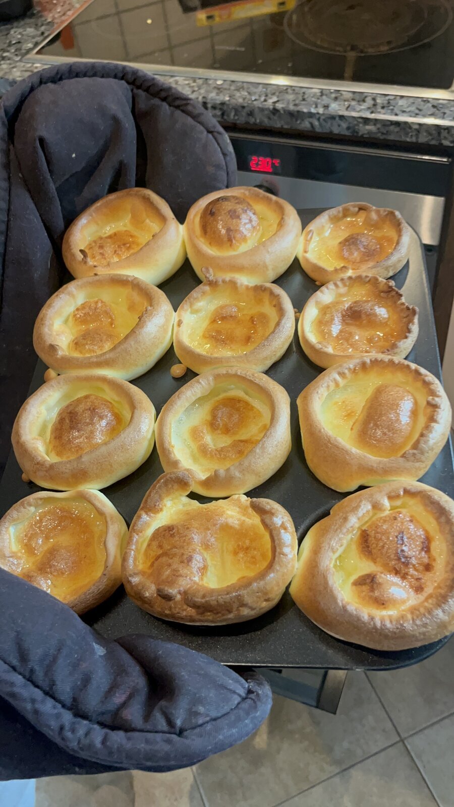 A 12-cup tray of risen Yorkshire puddings being lifted from an oven reading 230 degrees C