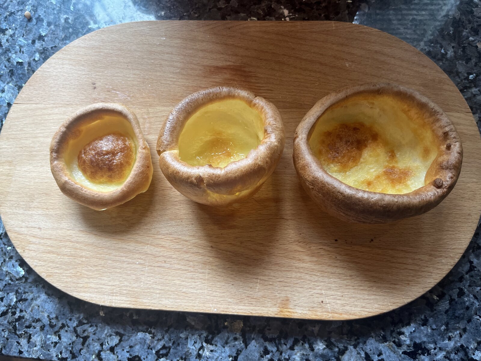 Three Yorkshire puddings of increasing size on a wooden board: a small one from the 12-cup tray, a medium from the 4-cup tray, and a large deep one from the jumbo muffin tray