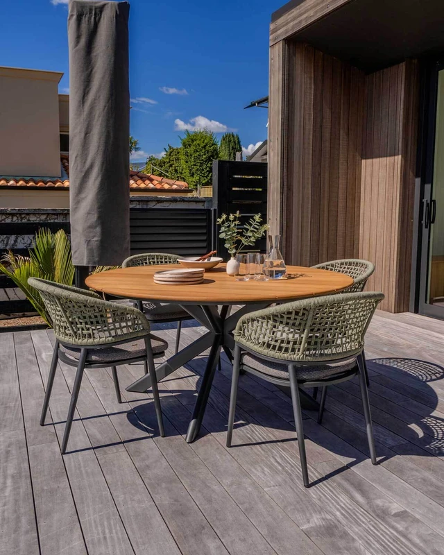 Paragon round outdoor dining table with teak timber top, paired with Noah woven outdoor dining chairs on a timber deck.