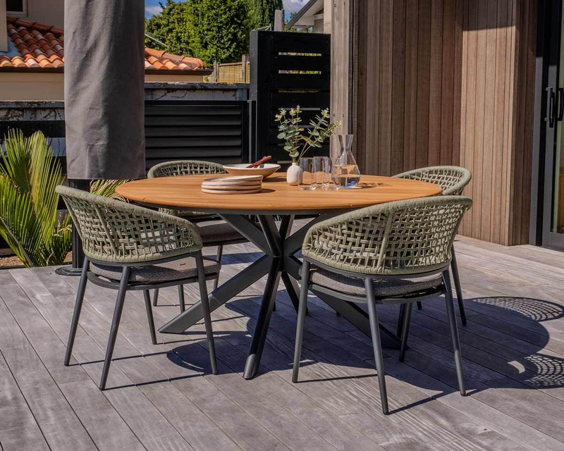 Paragon round outdoor dining table with teak timber top and charcoal geometric base, paired with Noah woven outdoor dining chairs on a timber deck.
