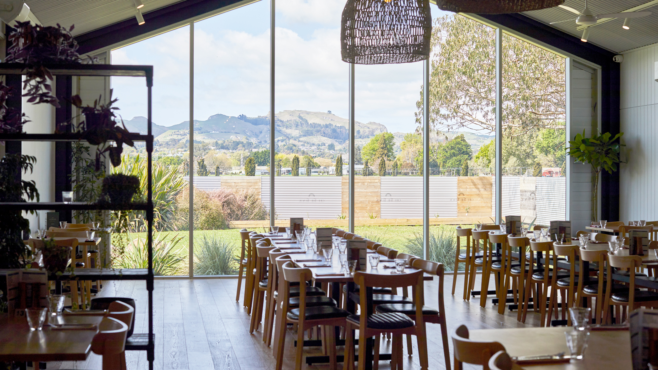 A modern restaurant interior with wooden restaurant tables and chairs set with plates and glasses, green cushioned bench seating, bar stools at the counter, and a single person sitting near the window.