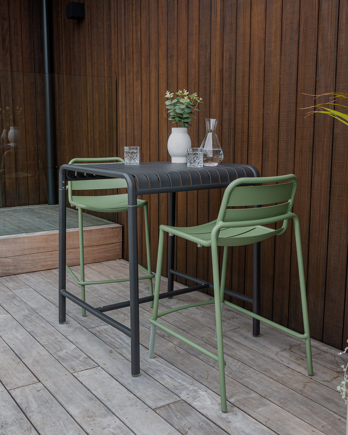 Atticua charcoal high table paired with a soft green Frankie bar stool in front of a timber clad wall with greenery