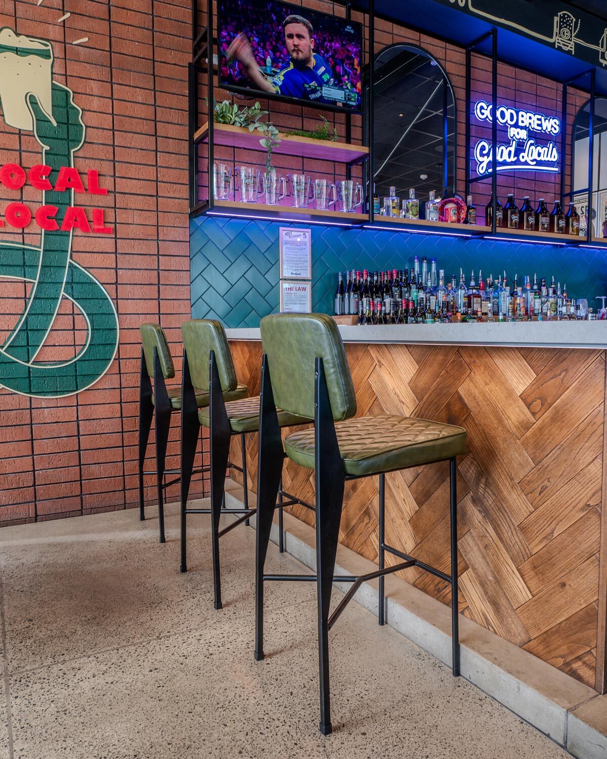 Dave upholstered bar stools styled along a hospitality bar setting, featuring quilted olive upholstery, black metal frames, and warm timber detailing.