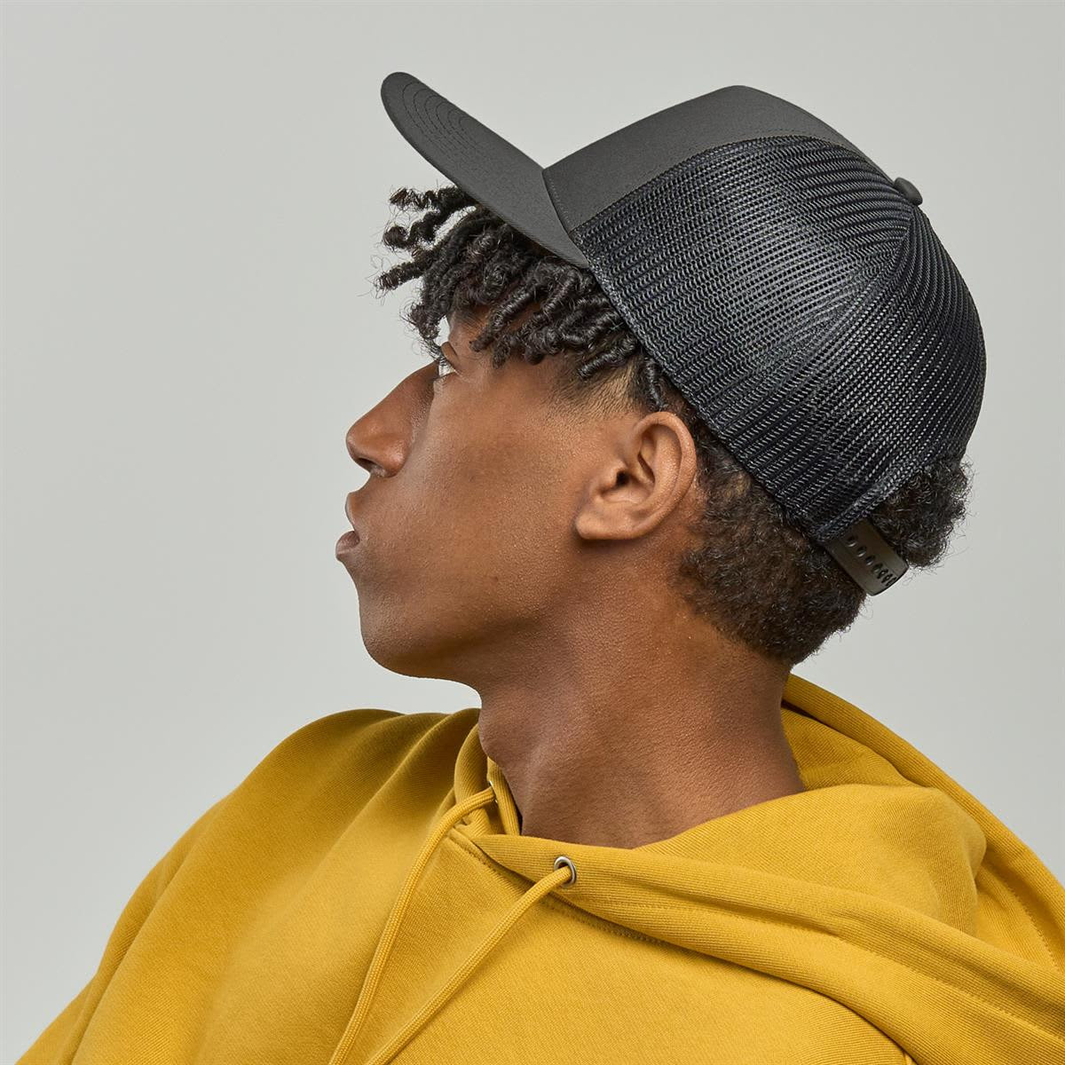 A black trucker cap with a mesh back, worn by a person in a yellow hoodie, facing sideways.