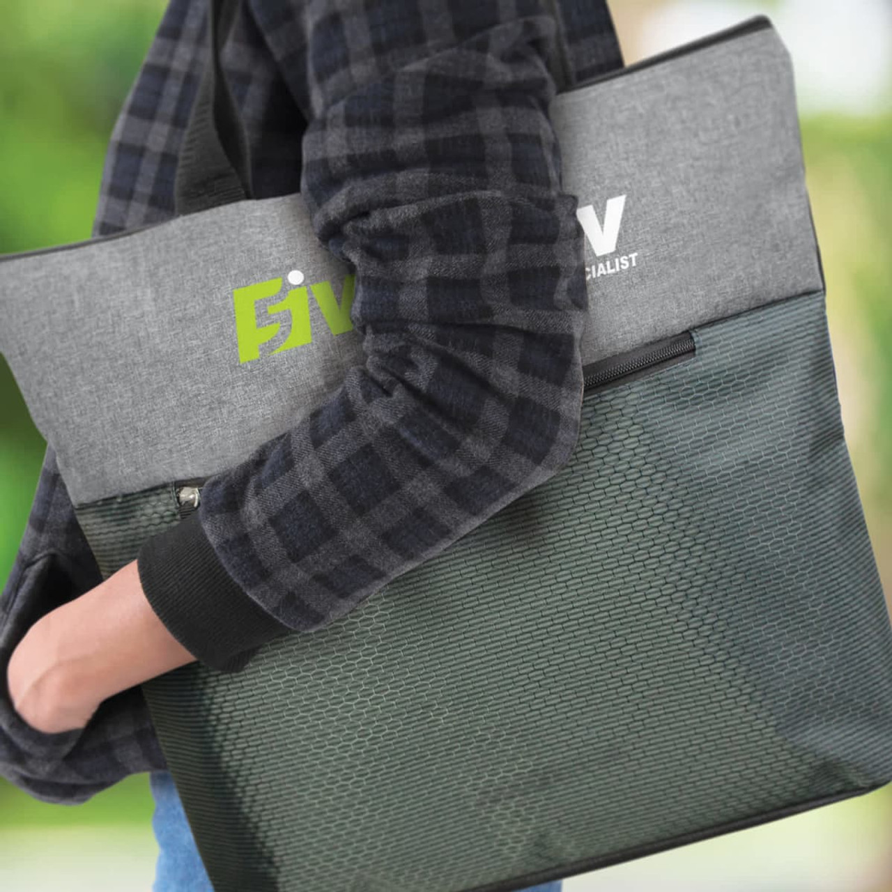 A grey and green tote bag is held by a person in a checkered jacket. The bag has a logo on it.