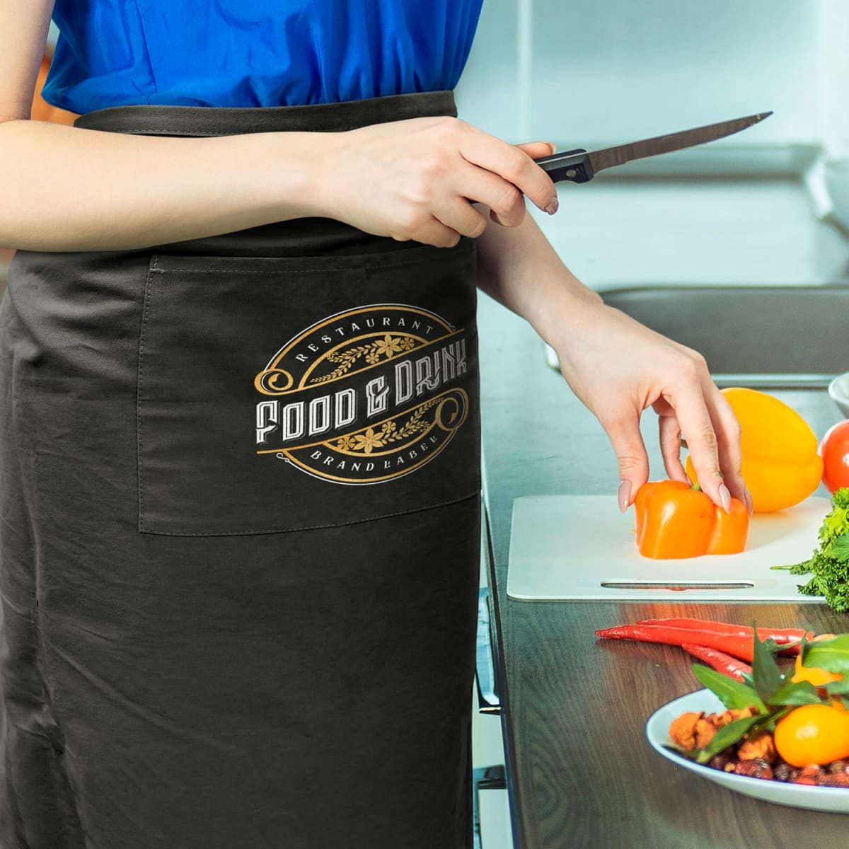 A black waist apron with a logo, worn by a person slicing bell peppers on a kitchen counter.