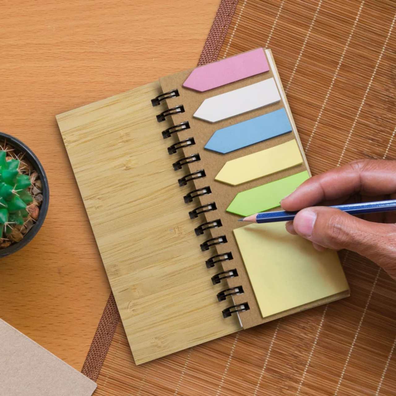 A bamboo sticky note wallet featuring sticky note flags in pink, blue, yellow, and green, alongside a pen and a small plant.