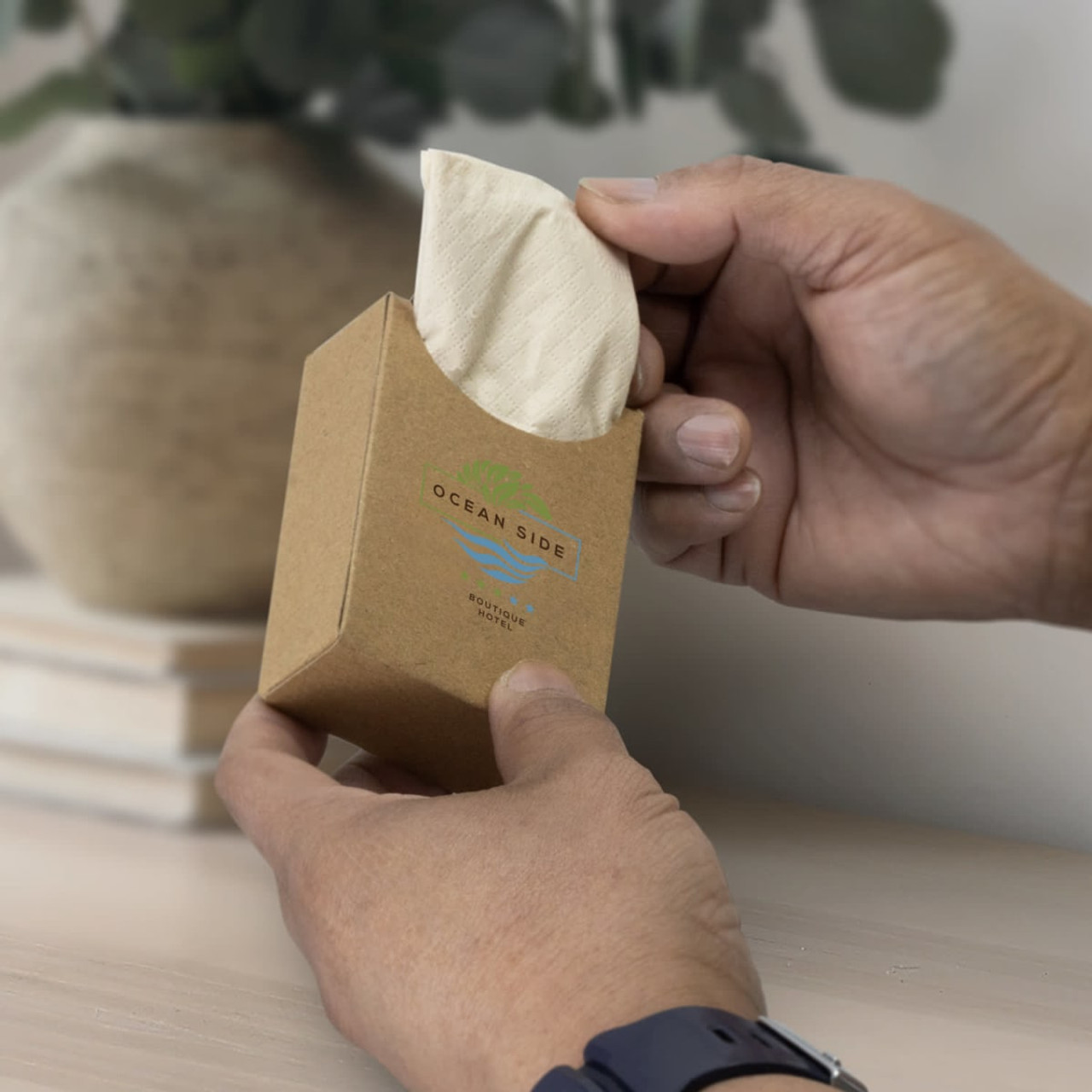 A hand holds a rectangular, brown kraft box containing travel tissues with a textured, light-coloured tissue visible.