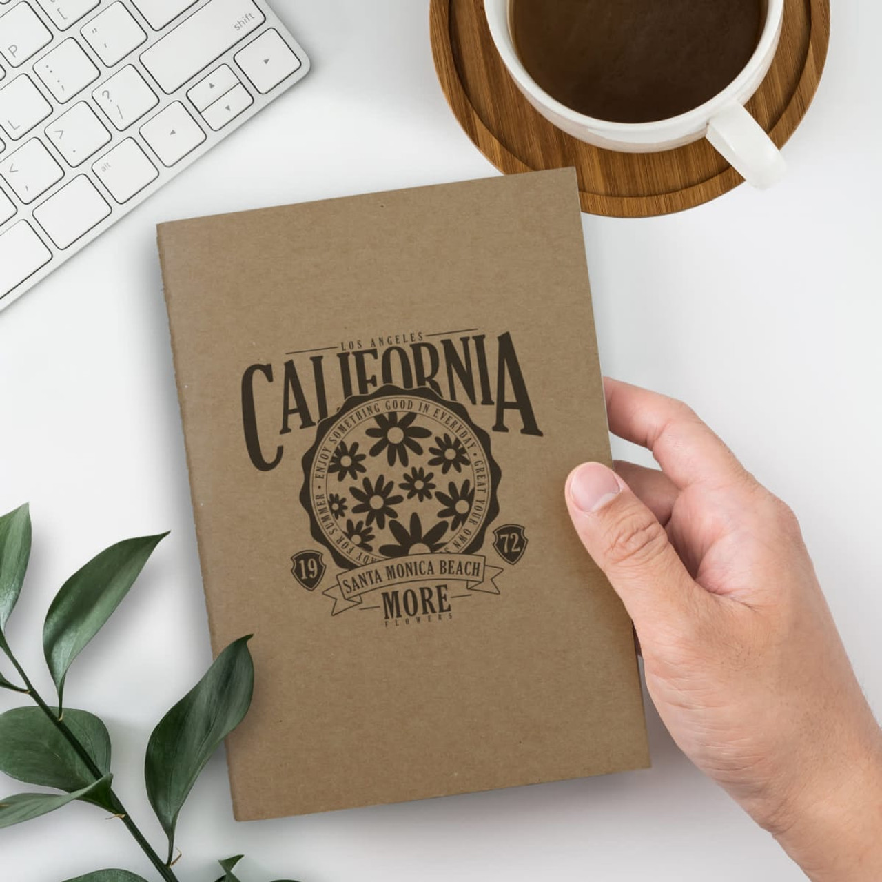 A brown soft cover notebook with a floral design, held in one hand, next to a coffee cup and keyboard.