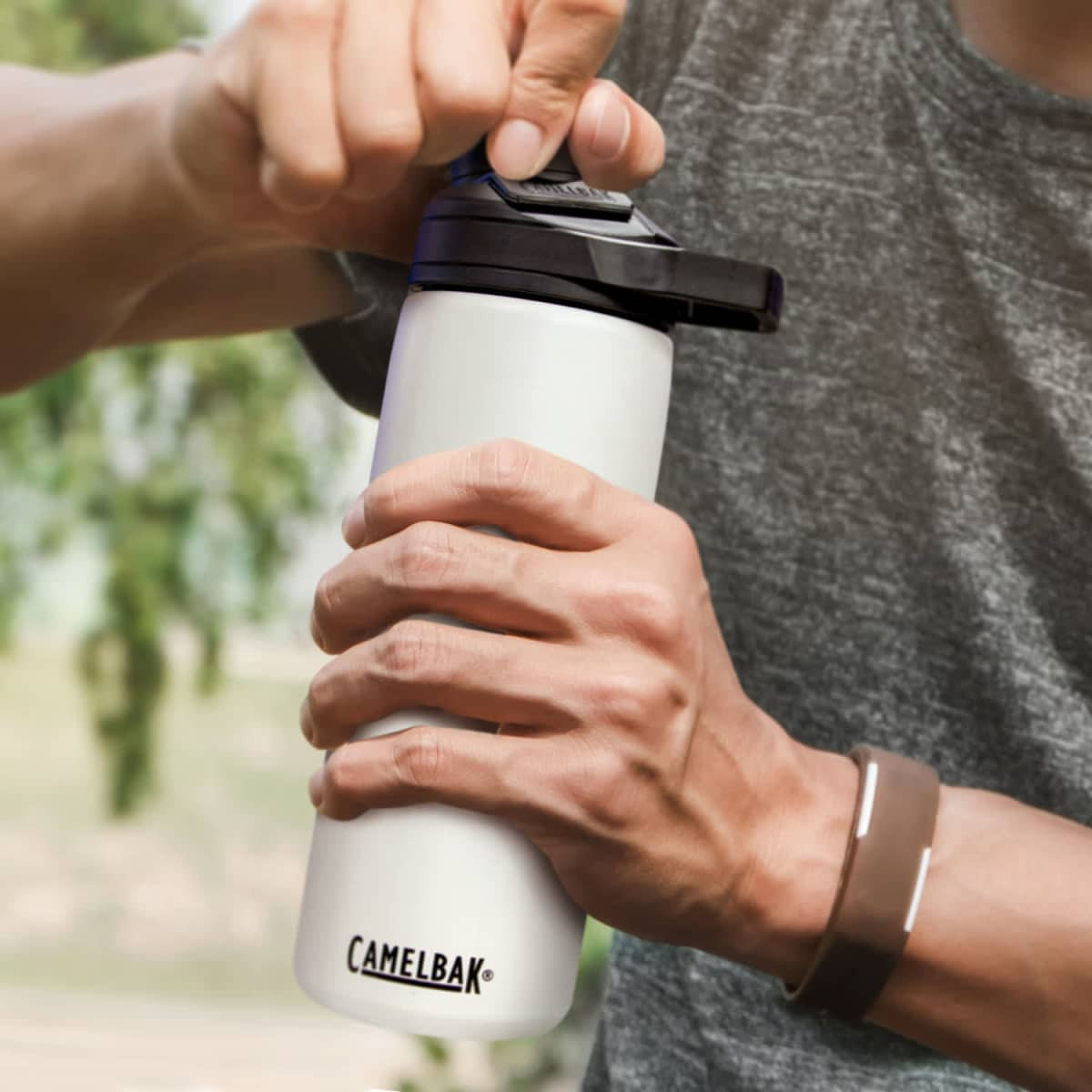 A drink bottle in white with a black lid, featuring a logo, being held and operated by a person.