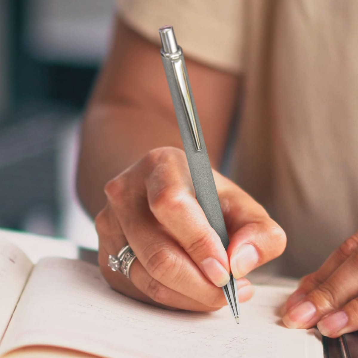 A metal pen with a sleek grey finish, held by a person writing in a notebook. Features a logo on the pen.
