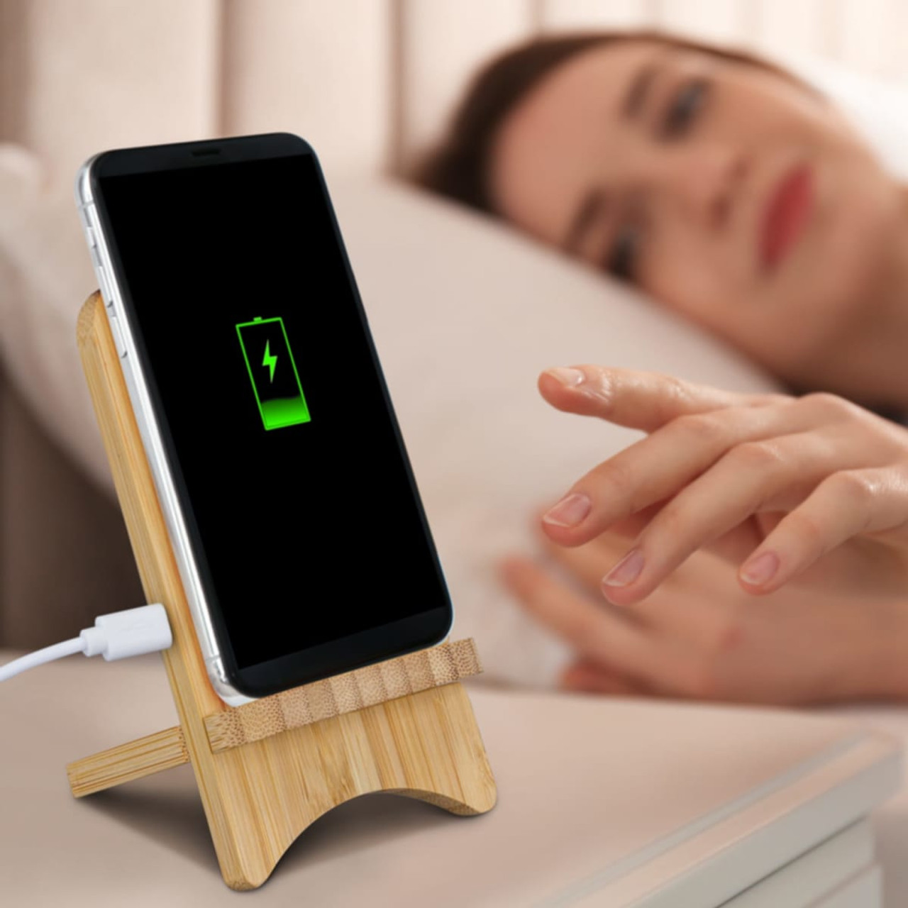 A bamboo wireless charging stand holds a smartphone displaying a battery icon, set on a bedside table.