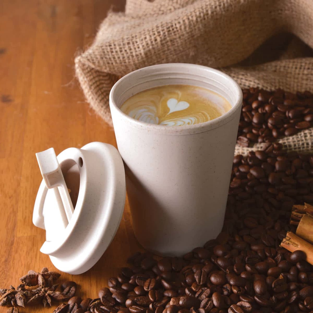 A reusable coffee cup in light beige with an eco-friendly lid, surrounded by coffee beans and burlap.