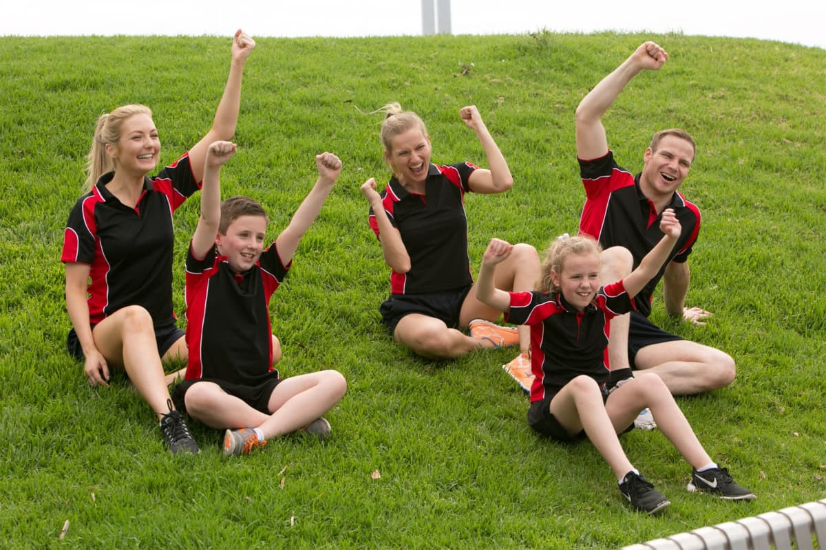 A group of five people wearing black and red polo shirts, celebrating and posing joyfully on green grass.