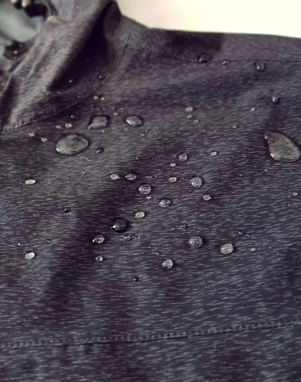 A close-up of a black waterproof performance jacket featuring droplets of water on the surface.