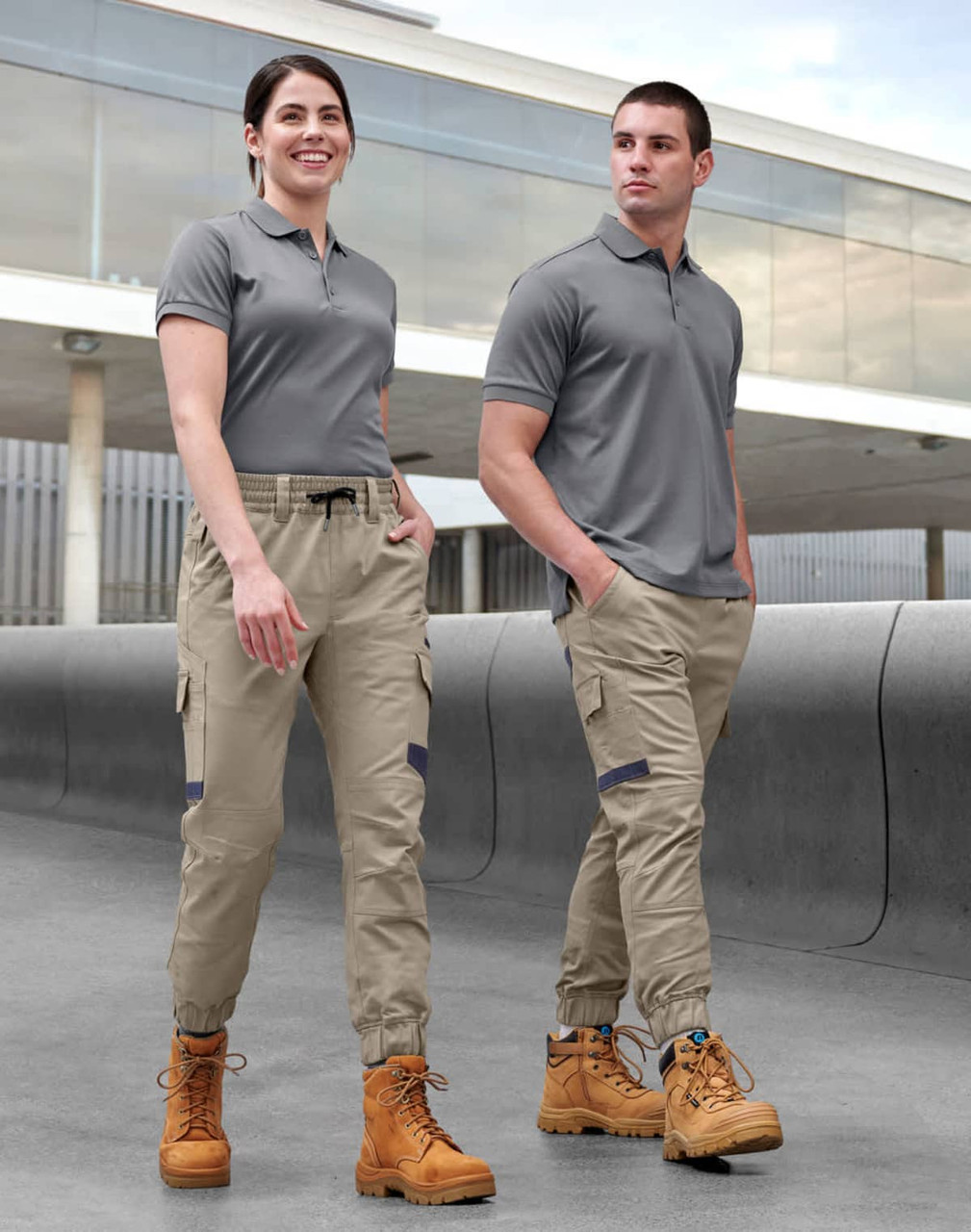 Unisex cotton stretch drill cuffed work pants in khaki, modelled by two people wearing grey tops and work boots.