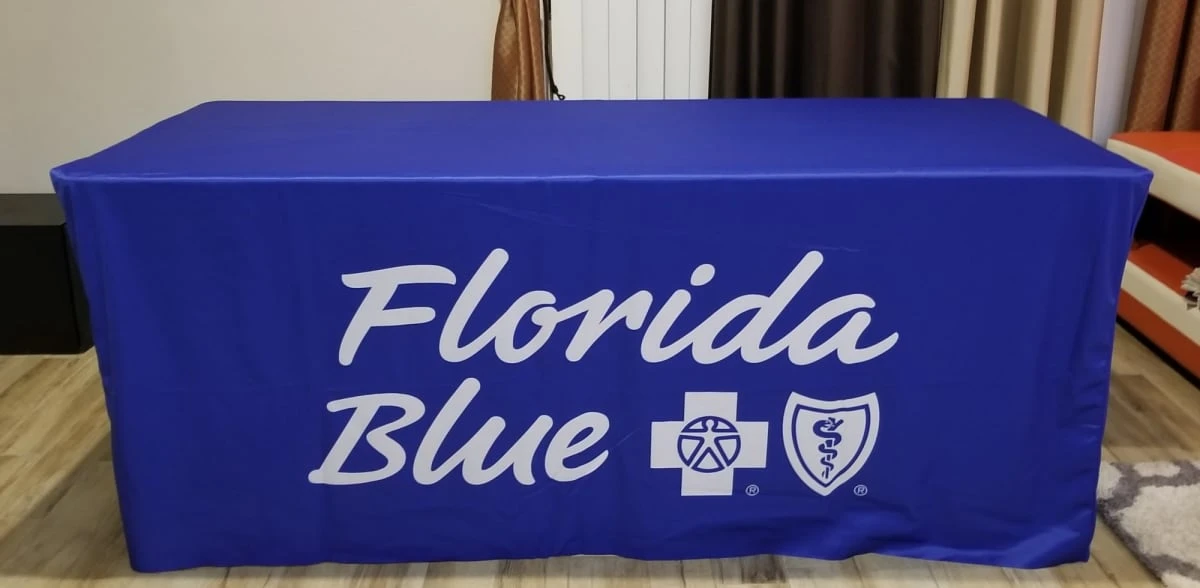 A blue fitted table cover with a logo and white text. Suitable for an 8-foot table display.