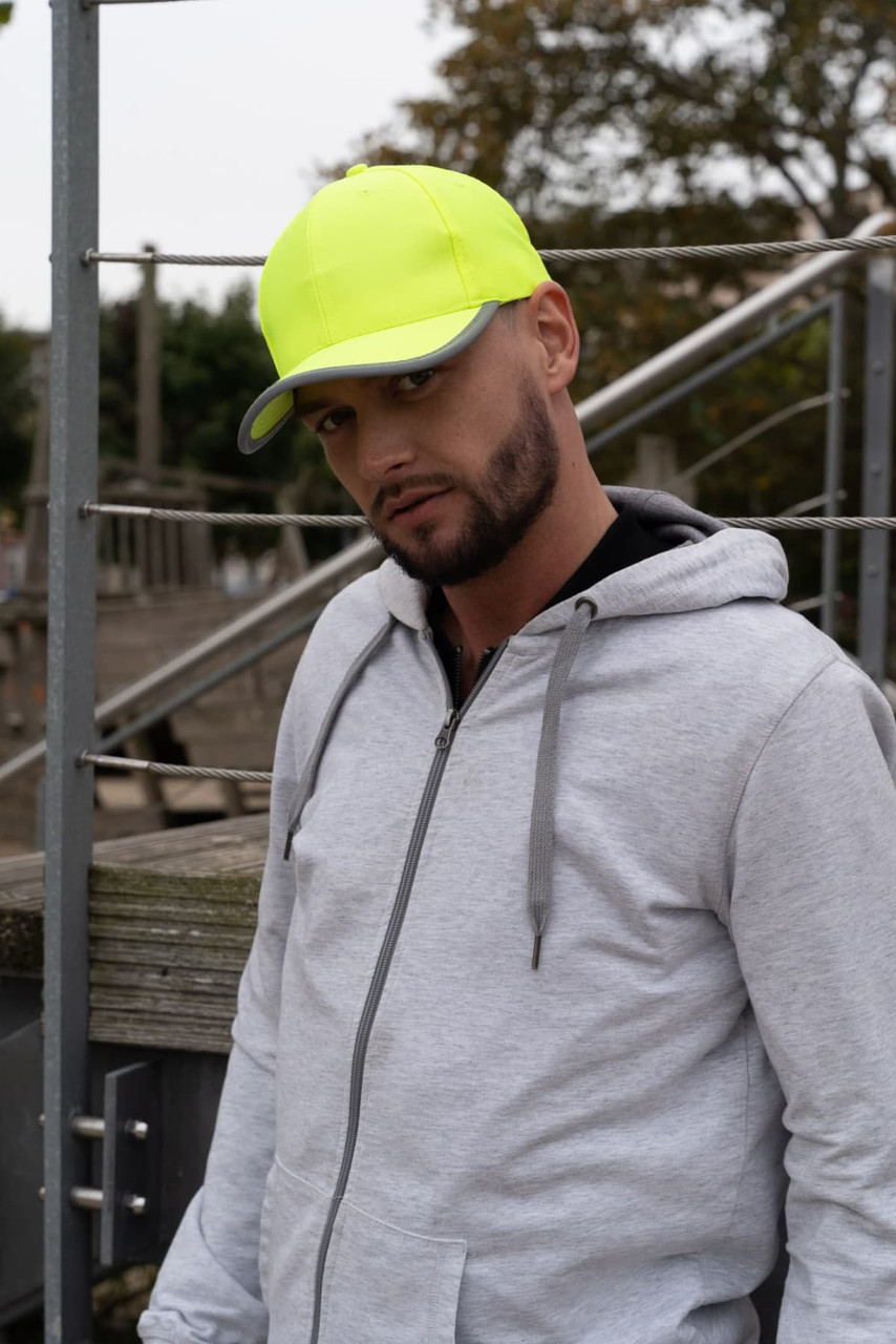 A man wearing a bright yellow cap stands outdoors, paired with a grey hoodie. The cap features reflective trim and a logo.