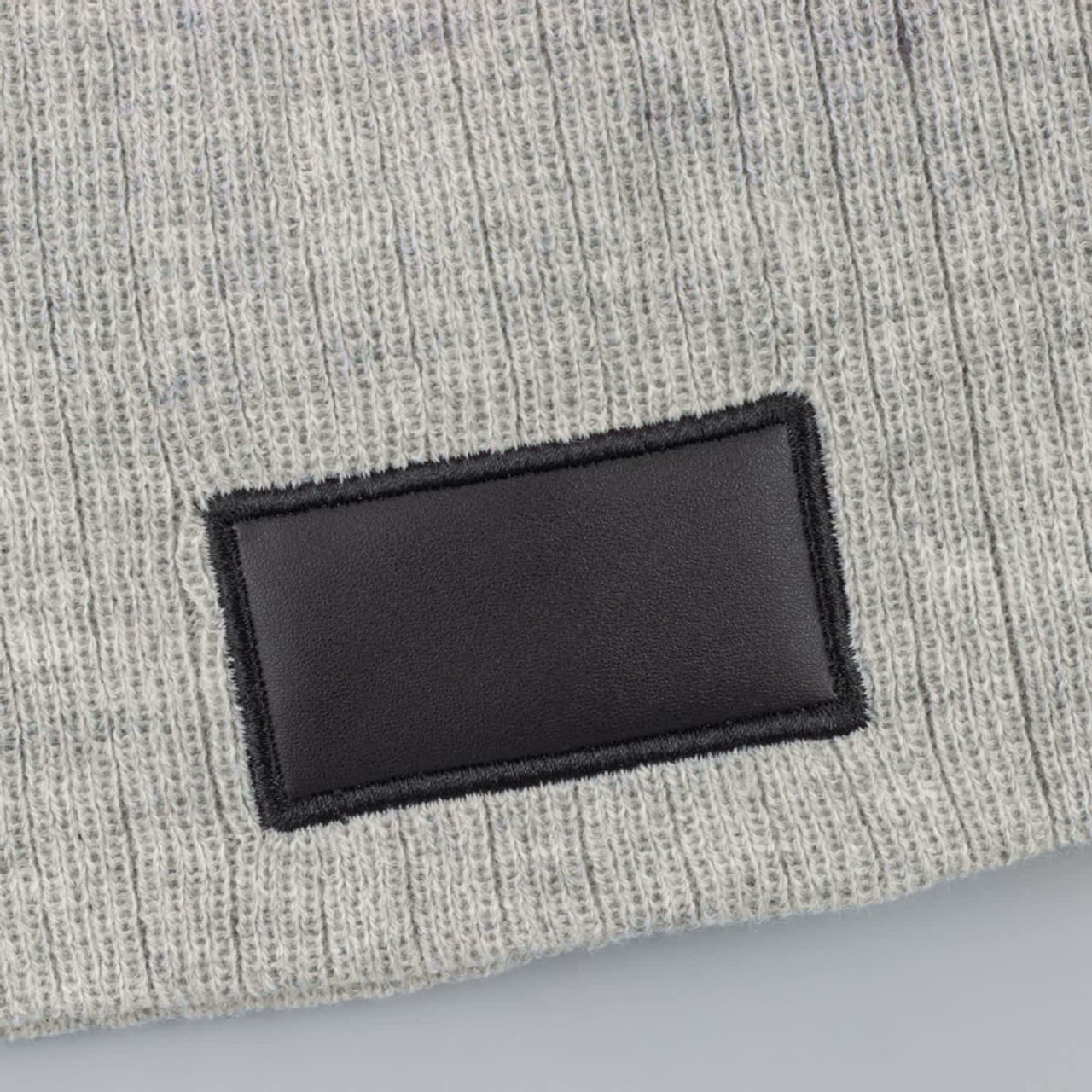 A grey cable knit beanie featuring a black patch, made from soft wool blend material.