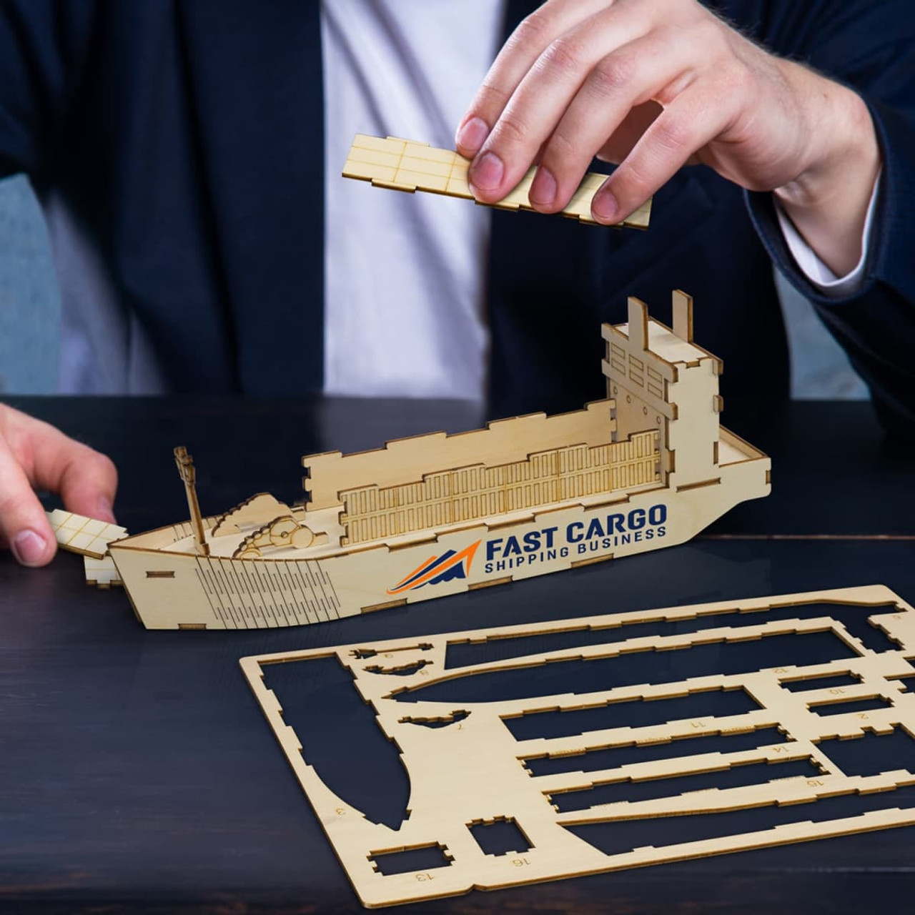 A wooden model of a cargo ship with a person assembling parts, featuring natural wood tones and a logo.