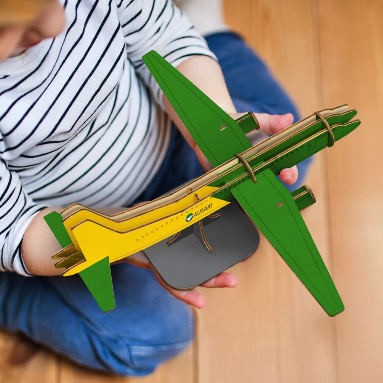 A wooden model of a jet plane in green and yellow, held by a child. The model is mounted on a grey base.