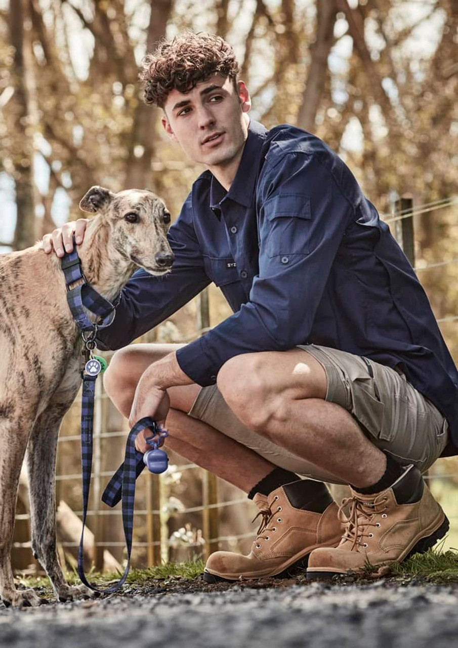 A man in a navy long sleeve button-up shirt kneels next to a greyhound on a leash. The background features trees.