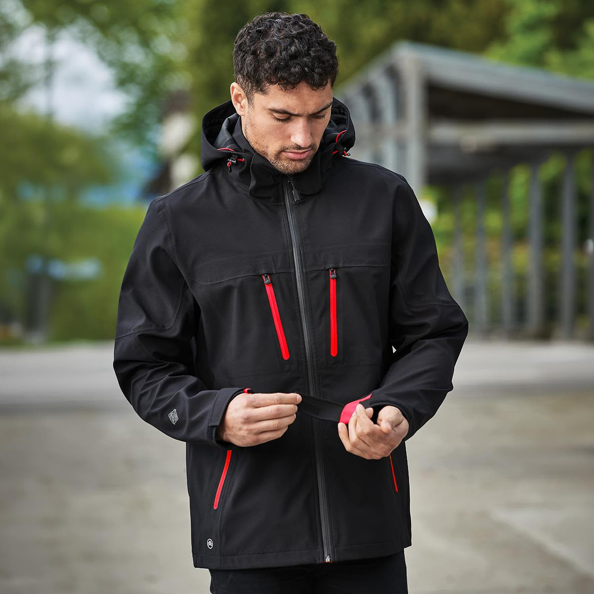 A black softshell jacket with red zippers and a hood, featuring a logo on one side.