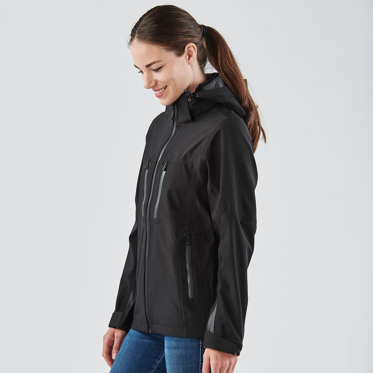 A woman wearing a black softshell jacket with a hood, featuring zippered pockets and a fitted design.