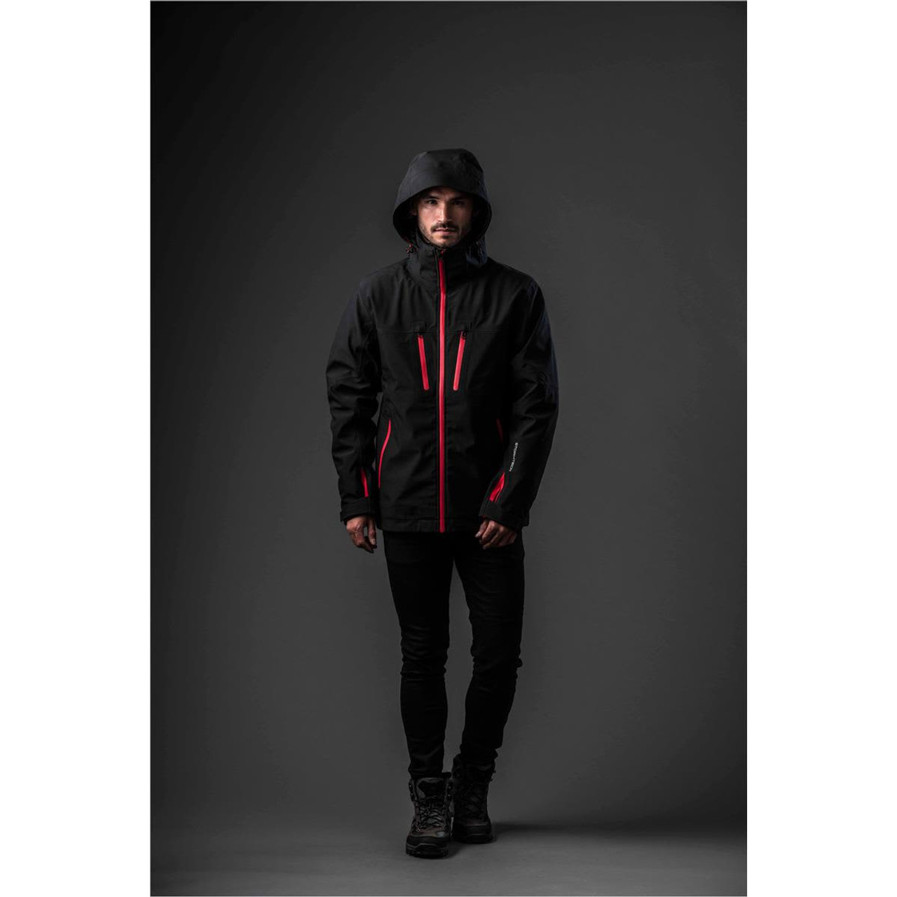 A man stands wearing a black jacket with red accents and a hood, featuring a branding logo.