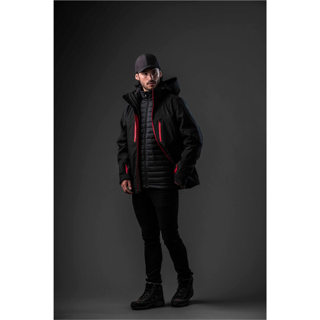 A man in a black jacket with red accents layered over a quilted jacket, wearing a cap and dark pants.