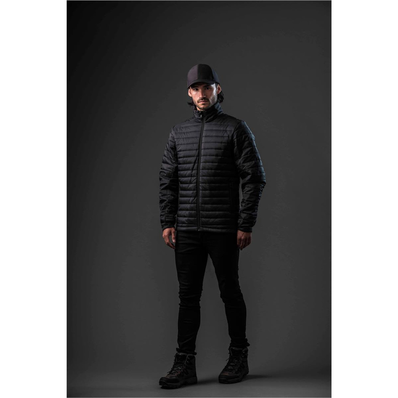 A man wearing a black puffer jacket stands against a dark background, complemented by black trousers and boots.