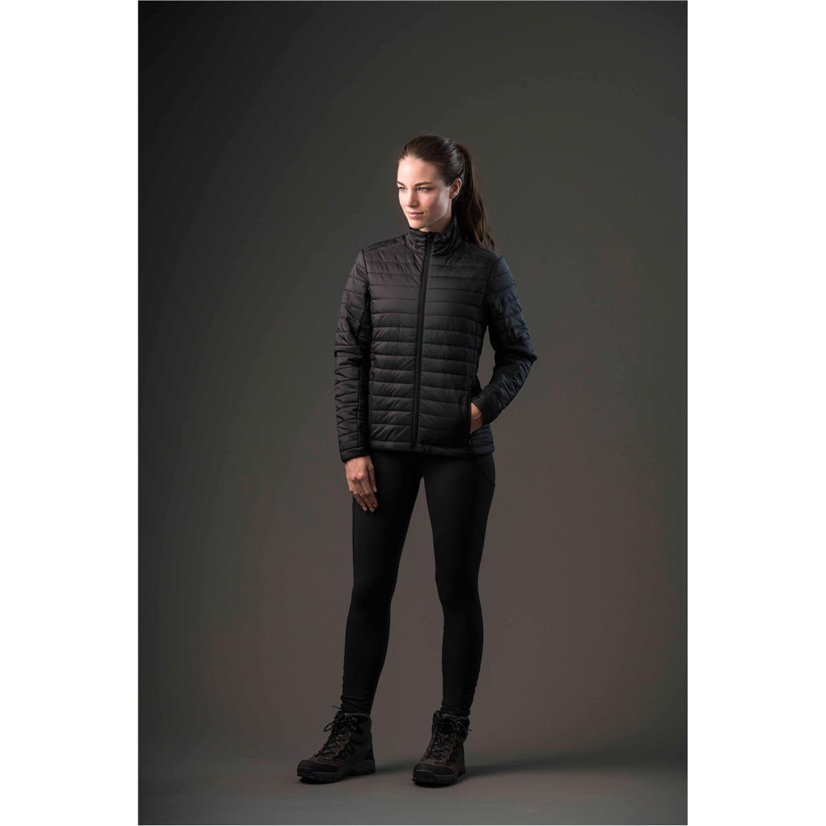 A woman models a black puffy jacket, paired with black leggings and boots, set against a dark background.